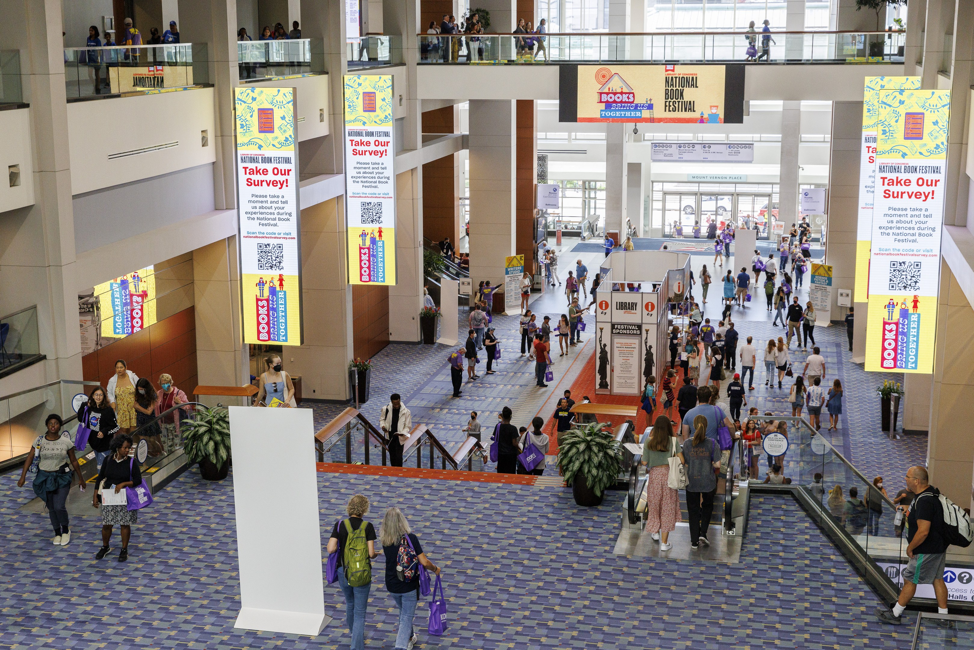 Eine große Menschenmenge, die durch ein großes Gebäude mit Bannern, Topfpflanzen, Rolltreppen, Geländern, Säulen, Texttafeln, Glastüren und anderen Gegenständen geht, wobei einige Taschen tragen.