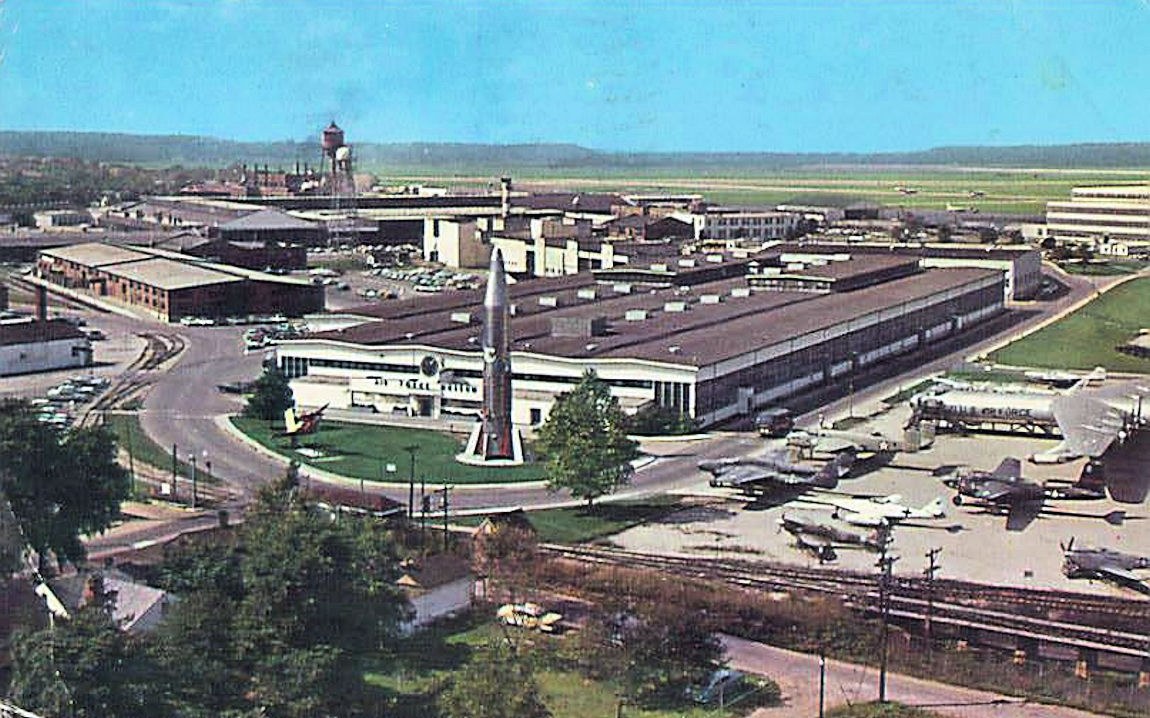 Altes Schwarz-Weiß-Foto eines Flughafens mit parkenden Flugzeugen, Gebäuden, Bäumen, Straßen, Fahrzeugen, Strommasten, Gras, Bahnschienen, Türmen, Hügeln und einem klaren blauen Himmel, mit Text unten.
