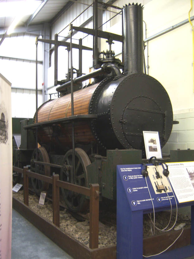 Alte Dampflokomotive in einem Museum mit Informationsschildern, einem Zaun, Steinboden, einer Fahne, einer Wand mit Rohren und Deckenbeleuchtung ausgestellt.