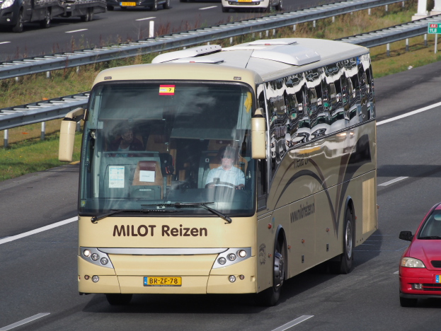 Ein Minibus mit Passagieren fährt auf einer Autobahn neben einem roten Auto, mit Geländern, Gras und textbehafteten Säulen im Hintergrund.