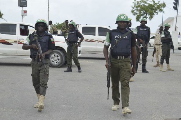 Gruppe von Polizisten in Helmen und Uniformen, die eine Straße entlanggehen und Waffen tragen, mit Fahrzeugen, Verkehrszeichen, Bäumen, Gebäuden und einem bewölkten Himmel im Hintergrund.