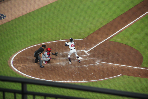 Ein Baseballspieler in einem Sportdress schwingt einen Schläger auf einem grünen Feld, wobei ein Geländer am unteren Bildrand zu sehen ist.