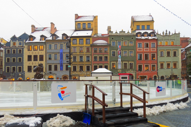 Eislaufbahn in einem Stadtplatz umgeben von Gebäuden, Treppen, Geländern, Informationsschildern, Laternen, Bäumen und schneebedecktem Boden unter einem Himmel.