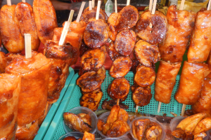 Marktstand mit Spießen von Hähnchenflügeln neben anderen Lebensmitteln in Plastikbehältern, mit Menschen und Flaschen im Hintergrund.