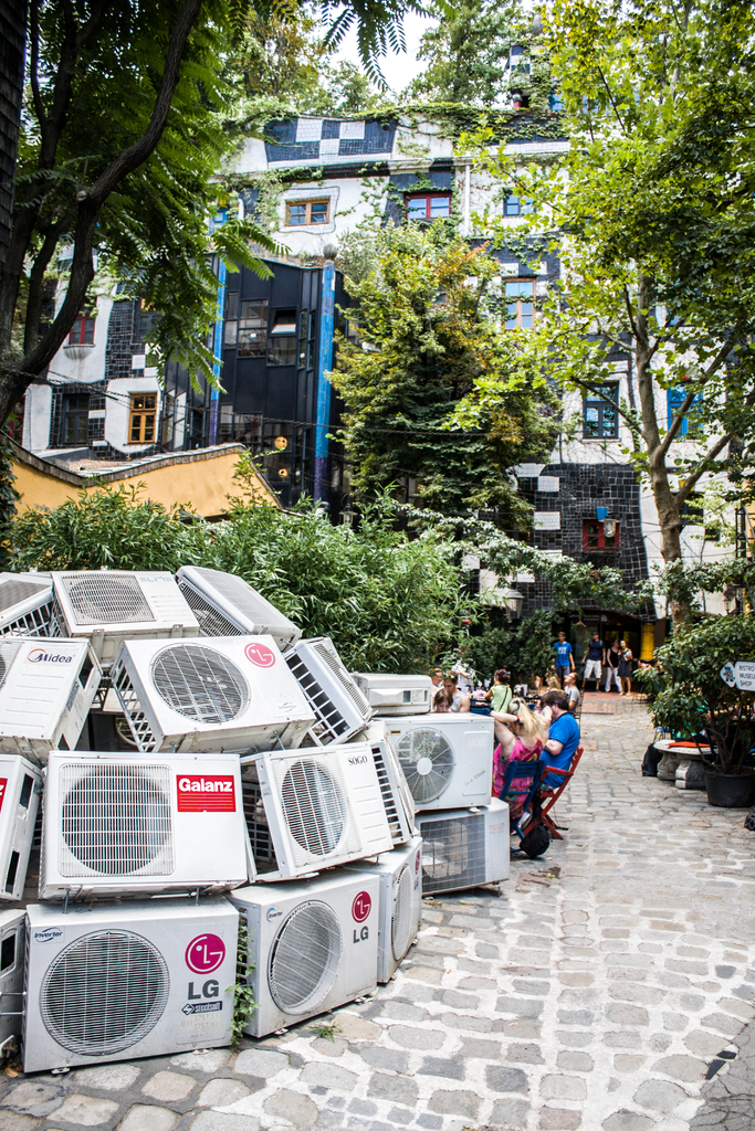 Luftkühler am Boden links unten, ein Weg rechts daneben, Menschen auf Stühlen neben dem Weg sitzen, mit Gebäuden, Bäumen und Pflanzen oben.