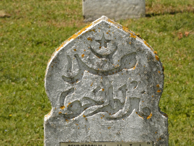 Ein steinerner Grabstein mit arabischer Schrift in einem grasbewachsenen Friedhof.