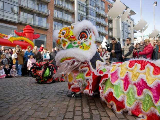 Ein lebendiges chinesisches Neujahrsfest in Amsterdam mit einer Löwendance-Aufführung vor einer Zuschauermenge, einige halten Kameras, vor einem Hintergrund aus Gebäuden, Laternenmästen und einem klaren blauen Himmel.