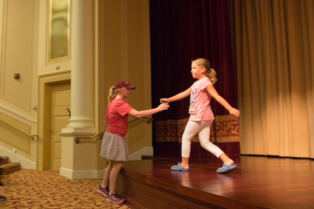 Zwei junge Mädchen tanzen auf einer Bühne vor einem Vorhang, mit einer Person, die auf einem Stuhl zu ihrer linken Seite sitzt, einer Säule und einer Wand im Hintergrund.