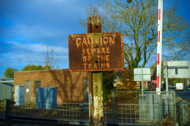 Vorsichtsschild auf einer Bahnschiene umgeben von einem Metallzaun, Bäumen, Strommasten, einem Gebäude mit Fenstern, Containern, einem Straßenmast, Fahrzeugen auf der Straße und einem bewölkten Himmel.