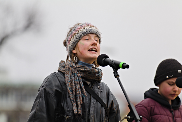 Eine Frau in schwarzer Jacke, Schal und Mütze singt in ein Mikrofon auf der Bühne, neben einer Person in einer maronenfarbenen Jacke und schwarzer Mütze, während ein unscharfer Hintergrund die Menge bildet.
