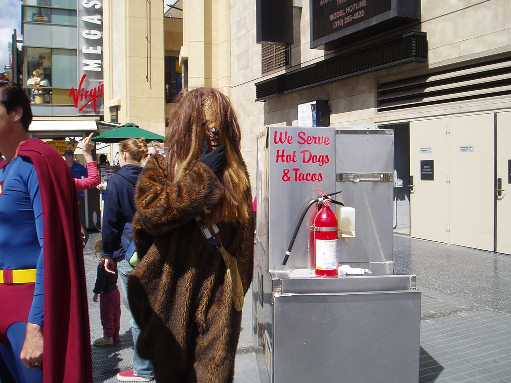 Eine Menschenmenge, einige in Kostümen, versammelte sich vor Gebäuden mit Schildern, einer Box mit Schrift und einem Feuerlöscher.