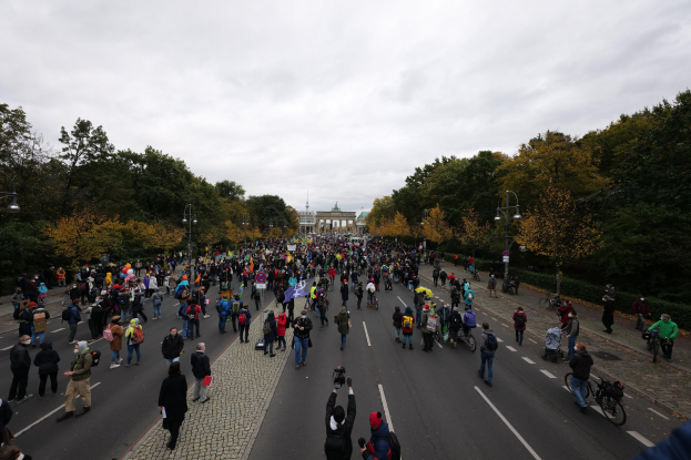 Eine große Gruppe von Menschen marschiert auf einer von Bäumen gesäumten Straße in Berlin, mit Kameras in der Hand, vor einem Gebäude und einem klaren Himmel.