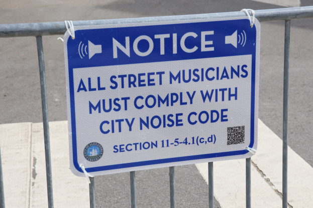 Hinweistafel an einem Zaun mit einem Schild über Straßenmusiker-Lärmvorschriften, mit einer Straße im Hintergrund.