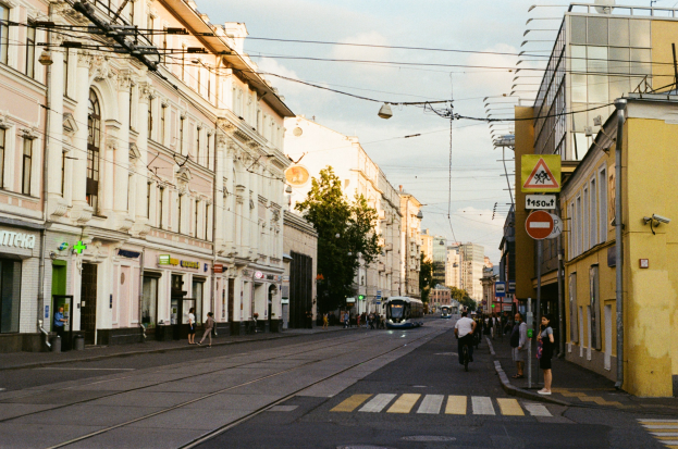 Eine Stadtstraße mit Fußgängern und Radfahrern, gesäumt von Gebäuden, Schildern, Bäumen und Strommasten.