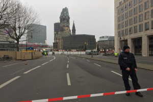 Polizist in schwarzer Uniform und Mütze auf einer Straße in Berlin umgeben von Verkehrskegeln, Fahrrädern, Laternenmasten, Bäumen und Gebäuden mit einem Kirchturm im Hintergrund bei klarem blauem Himmel.