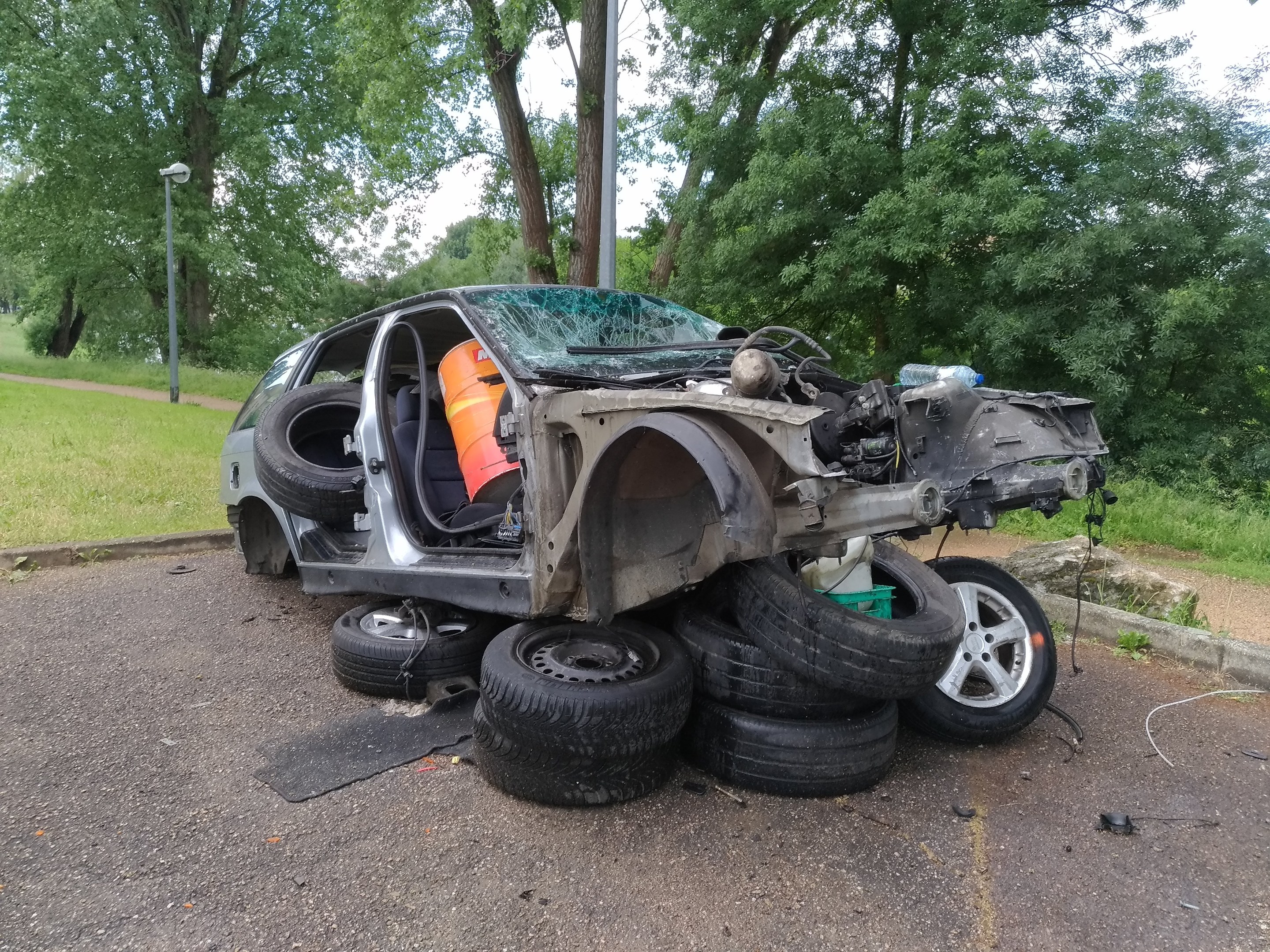 Ein umgestürzter Wagen am Straßenrand mit verstreuter Trägermaterial, umgeben von Gras, Pflanzen, Bäumen, einer Straßenlaterne und einem bewölkten Himmel.