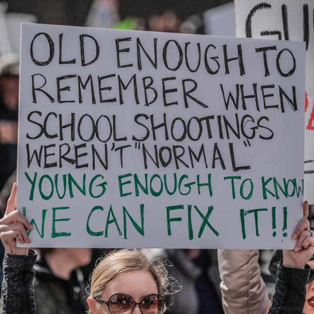 Eine Frau mit Schutzbrille hält ein Schild mit der Aufschrift "Alt genug, um sich an die Zeit zu erinnern, als Schulschießereien nicht normal waren, jung genug, um zu wissen, dass wir es ändern können" in der Hand, während im Hintergrund Gruppen von Menschen und ein unscharfer Hintergrund zu sehen sind.
