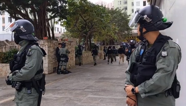 Eine Gruppe von Polizisten in Helmen und Schutzwesten steht auf einer Straße in Hong Kong, mit Bäumen, Gebäuden und Himmel im Hintergrund, whährend sie versuchen, Protestierende zu zerstreuen.
