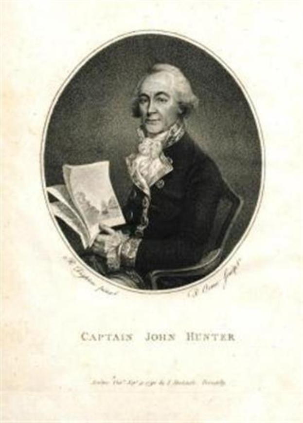 Captain John Hunter, der eine blaue Anzug, ein weißes Hemd und eine rote Krawatte trägt, sitzt in einem Stuhl und liest ein Buch mit einem ernsten Gesichtsausdruck, mit dem Text "Captain John Hunter" unten.