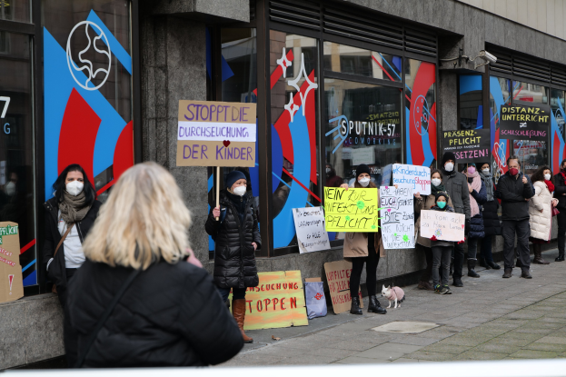 Gruppe von maskierten Menschen mit Schildern und Plakaten vor einem Glasfrontgebäude, mit Kameras und einem Hund, protestiert gegen ein deutsches Regierungsverbot von Masken in Schulen.