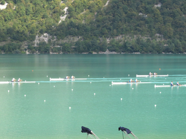 Gruppe von Menschen bei einem Ruderwettbewerb auf einem Gewässer mit Bäumen und einem Hügel im Hintergrund, Kleidung auf Ständen am Wasserrand.