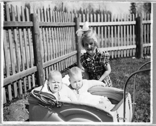 Schwarz-weißes Foto von drei Kindern in einem Auto, mit einem Mädchen, das im Vordergrund steht und lächelt, einem Zaun und Bäumen im Hintergrund und Gras auf dem Boden.