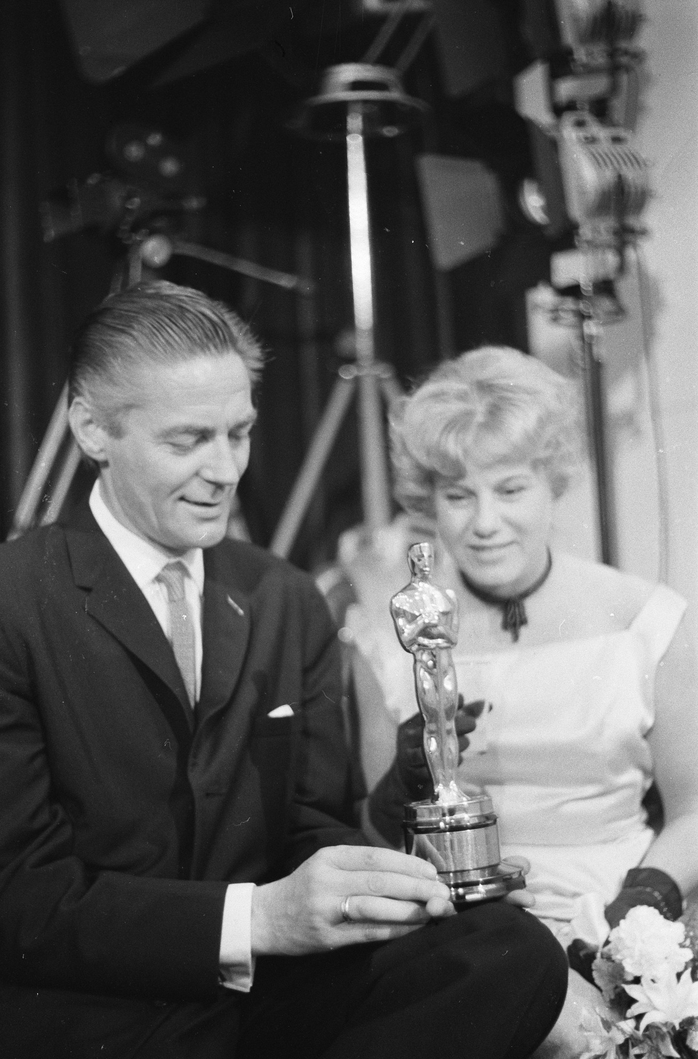 Ein Schwarz-Wei├č-Bild eines formell gekleideten Mannes mit einer Oscar-Statue und einer Frau mit einem Blumenstra├čus, die nebeneinander sitzen und von Hintergrundbeleuchtung umgeben sind.