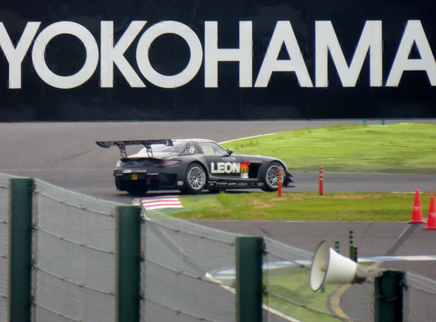 Schwarzer Sportwagen auf einer Rennstrecke mit Verkehrszeichen, umgeben von Gras und einem Zaun, mit einem "Yokohama"-Schild im Hintergrund und einem Lautsprecher im Vordergrund.