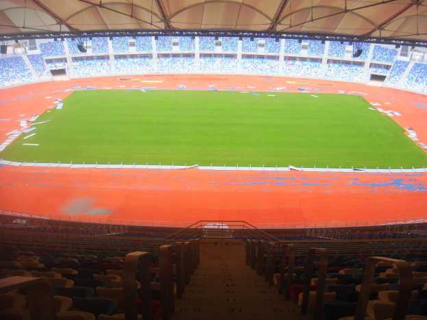 Großes Stadion mit vielen Zuschauern, mit Sitzreihen, Geländern, Treppen und Dach, mit dem Spielfeld in der Mitte sichtbar.