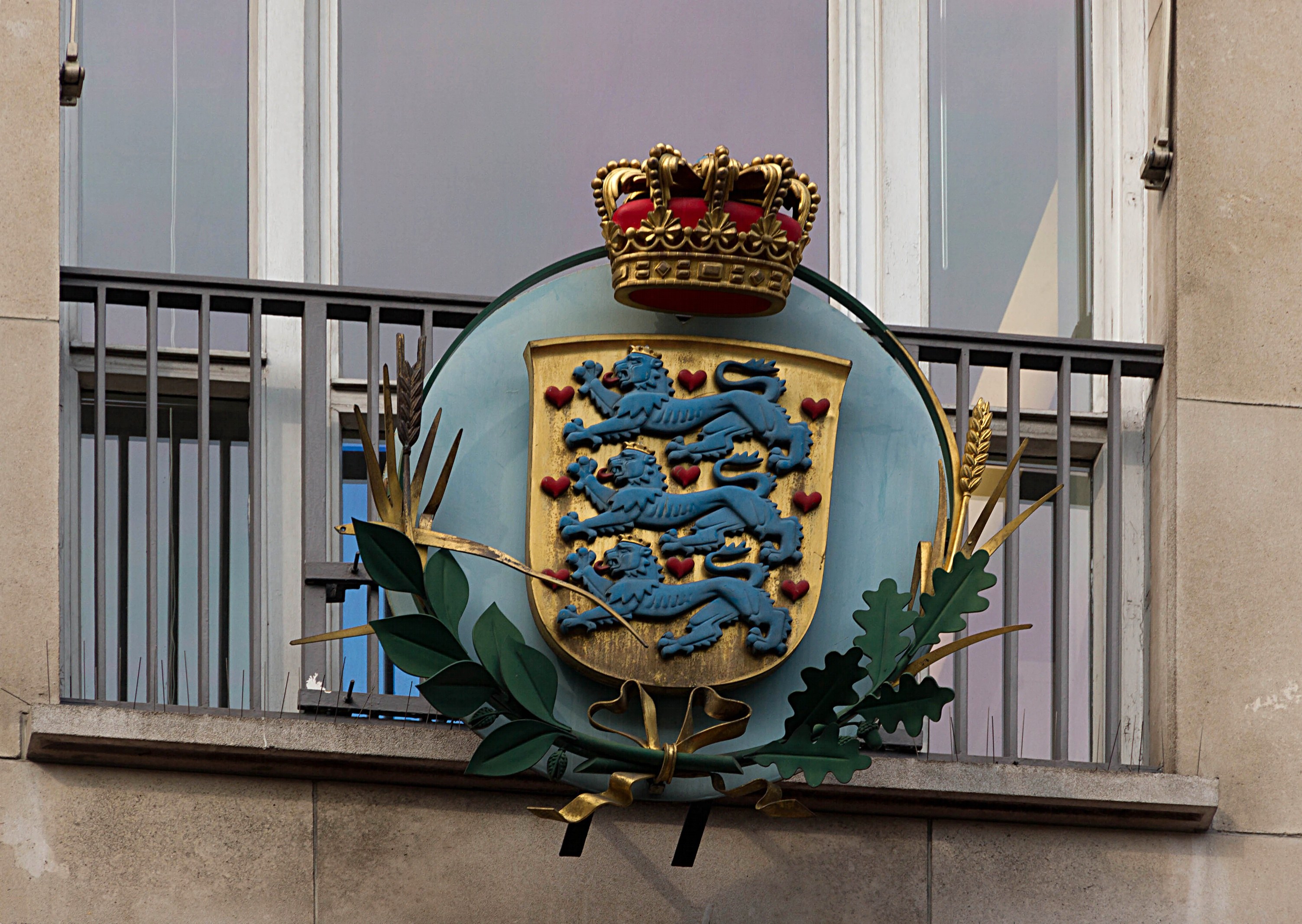 Das Bild zeigt das Wappen der britischen Königsfamilie auf einem Gebäude, begleitet von einem Kronen- und Blätterlogo, mit Fenstern und Gittern im Hintergrund.