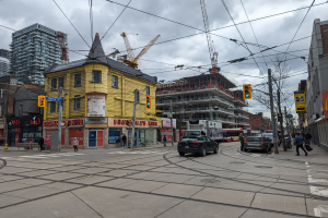 Stadtstraßenszene mit Autos, Fußgängern, Strommasten, Ampeln, Gebäuden, Schildern, Bäumen, Kränen und einem gelben Gebäude im Bau gegen einen bewölkten Himmel.