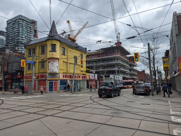 Stadtstraßenszene mit Autos, Fußgängern, Strommasten, Ampeln, Gebäuden, Schildern, Bäumen, Kränen und einem gelben Gebäude im Bau gegen einen bewölkten Himmel.