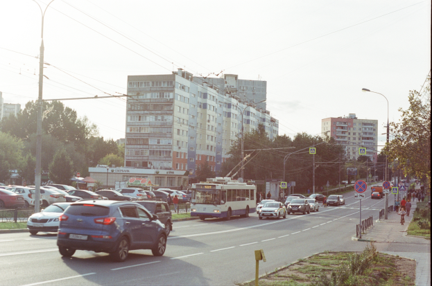 Eine belebte Stadtstraße mit zahlreichen Autos, Bussen, Straßenlaternen, Schildern, Laternenmasten, Strommasten mit Drähten, Bäumen und Gebäuden mit Fenstern unter einem klaren blauen Himmel.