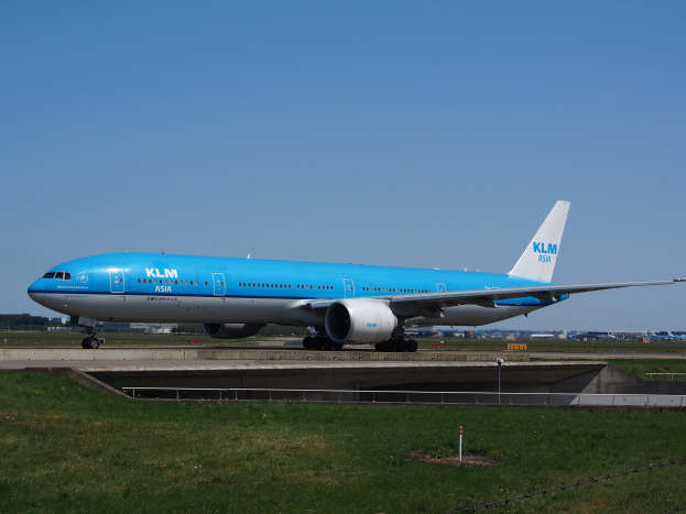 Ein KLM Boeing 777-300ER auf der Rollbahn am Amsterdam Airport, umgeben von Gras, einem Zaun, Pfählen, Gebäuden, Bäumen und einem klaren blauen Himmel.