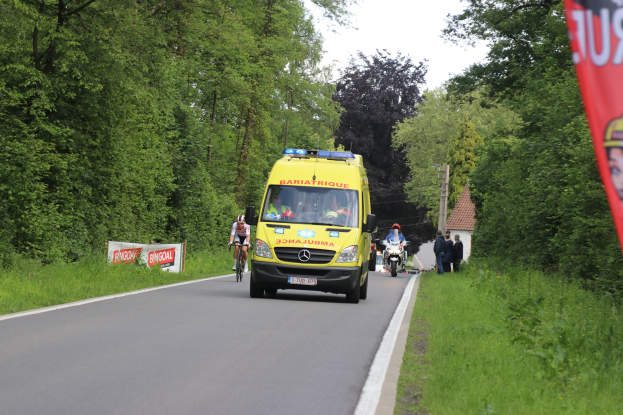 Ambulanz fährt auf einer Straße mit Radfahrern daneben, Gras und Bäume auf beiden Seiten, Häuser, Masten und einen klaren blauen Himmel im Hintergrund.
