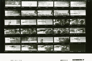 Ein Schwarz-Wei├č-Foto eines Mannes in einem wei├čem Hemd und Shorts, der einen Tennisschl├Ąger h├Ąlt und konzentriert in die Kamera schaut, mit einem Filmstreifen im Hintergrund.