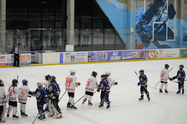Young people in helmets and hockey gear playing ice hockey on an indoor rink, with banners, pillars, and glass walls in the background.