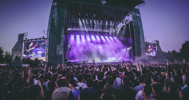 Eine große Menschenmenge schaut bei einem Musikfestival auf einer beleuchteten Bühne mit Instrumenten und Bildschirmen, die vor einem Gebäude, Bäumen und einem klaren blauen Himmel aufgebaut sind.
