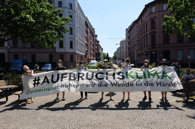 Maskierte Menschen halten ein Transparent mit der Aufschrift "Aufbruchsklima" während einer Klimademonstration in Berlin, umgeben von städtischen Elementen und anderen Teilnehmern.