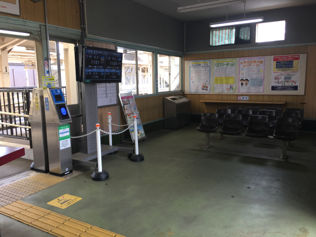 Ein belebter Bahnhofseingang mit Reihen von Stühlen, einem Service-Schalter, Ticketautomaten, Kettenzäunen, Informationsanzeigen, Wandplakaten, Deckenbeleuchtung und Geländern.