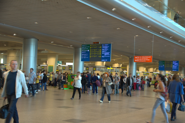 Ein volles Flughafen-Terminal mit Reisenden, die Gepäck tragen, übergeordnete Schilder, Deckenleuchten, Säulen und eine Glasbrüstung auf der rechten Seite.