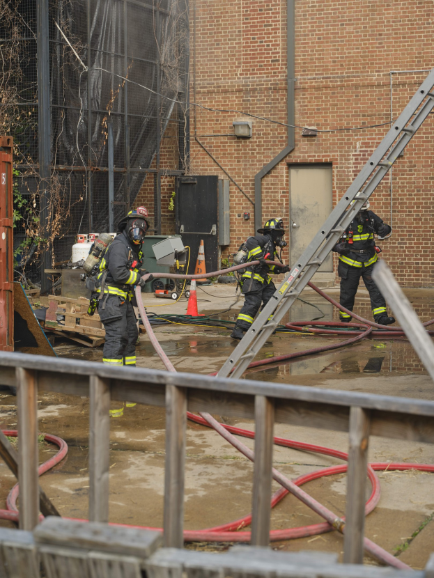Feuerwehrleute mit Helmen arbeiten daran, ein Gebäude Feuer zu löschen, umgeben von Ausrüstung wie Rohren, einem Metallzaun und einem Verkehrskegel, mit einem Baum und Himmel im Hintergrund.