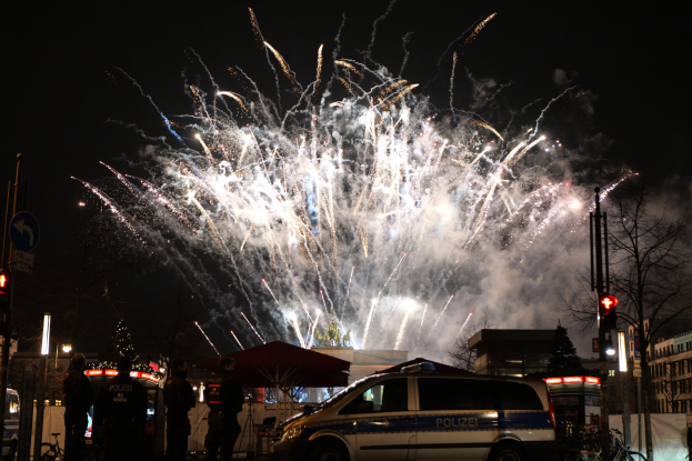 Polizeiauto vor einer Menschenmenge während einer Silvesterfeier in Berlin geparkt, mit farbenfrohen Feuerwerkskörpern am Himmel.