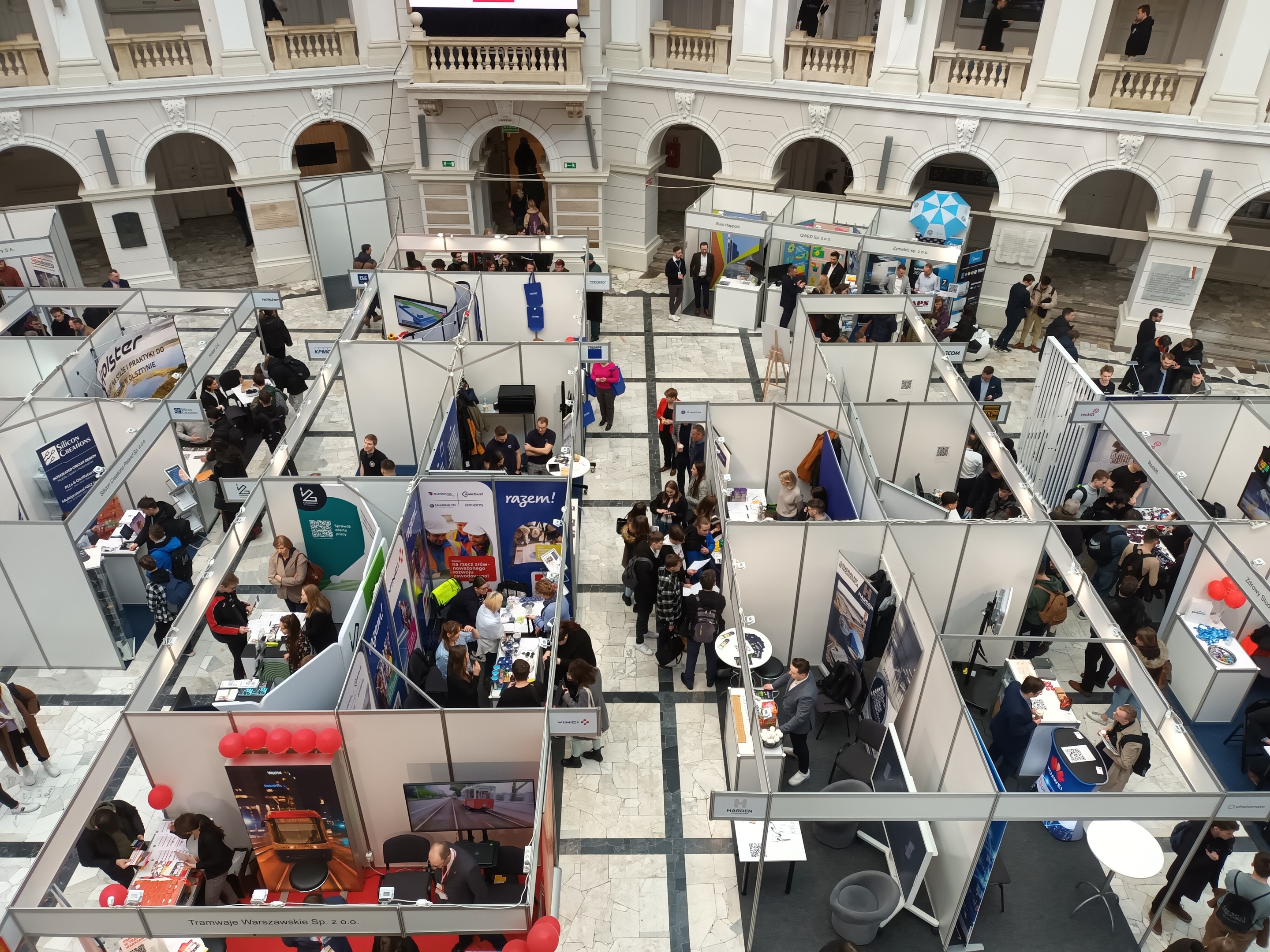 Große Gruppe von Teilnehmern auf einer internationalen Fachmesse mit Ständen, Bannern, Tischen, Stühlen und Luftballons in einem Raum mit architektonischen Bögen, Säulen und einem Bildschirm im Hintergrund.