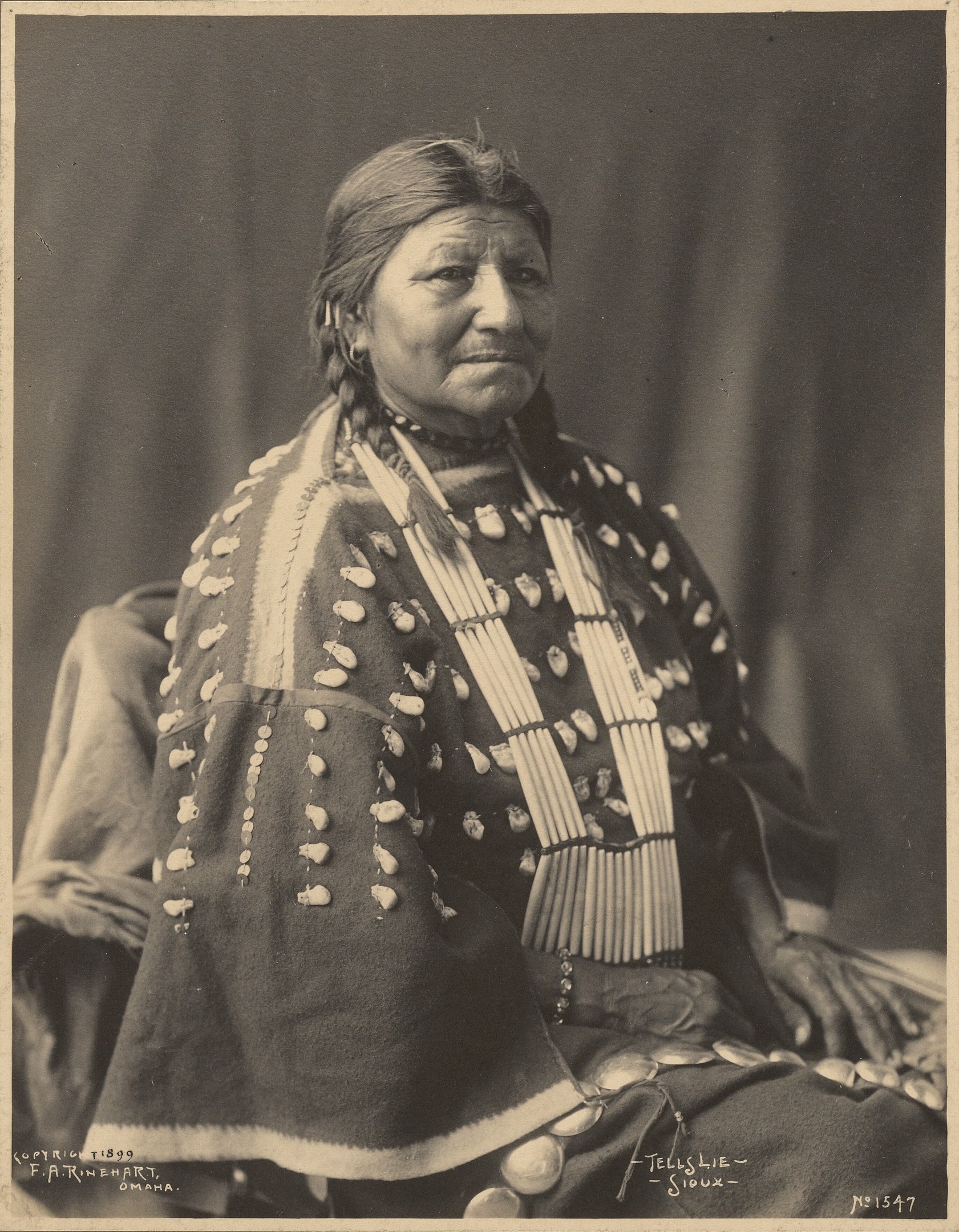 Altes Schwarz-Weiß-Foto einer indischen Frau in traditioneller Kleidung mit ernster Miene, gefalteten Händen, zurückgebundenem Haar und einem Federkopfschmuck.