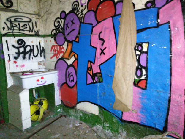 Ein Bad mit Wänden voller Graffiti, ein Waschbecken in der Ecke mit einem Tuch bedeckt und verstreute Gegenstände auf dem Boden.