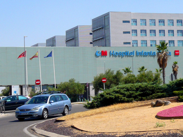 Krankenhaus mit parkenden Autos, umgeben von städtischen Elementen wie Straßenlaternen, Fahrzeugen, Grünflächen und einem klaren blauen Himmel.