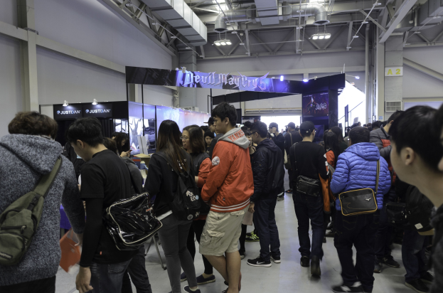 Eine Gruppe von Menschen vor einem vollen Gaming-Expo, mit einigen Besuchern mit Taschen, beleuchteten Schildern und an der Decke montierten Lampen und Rohren im Hintergrund.