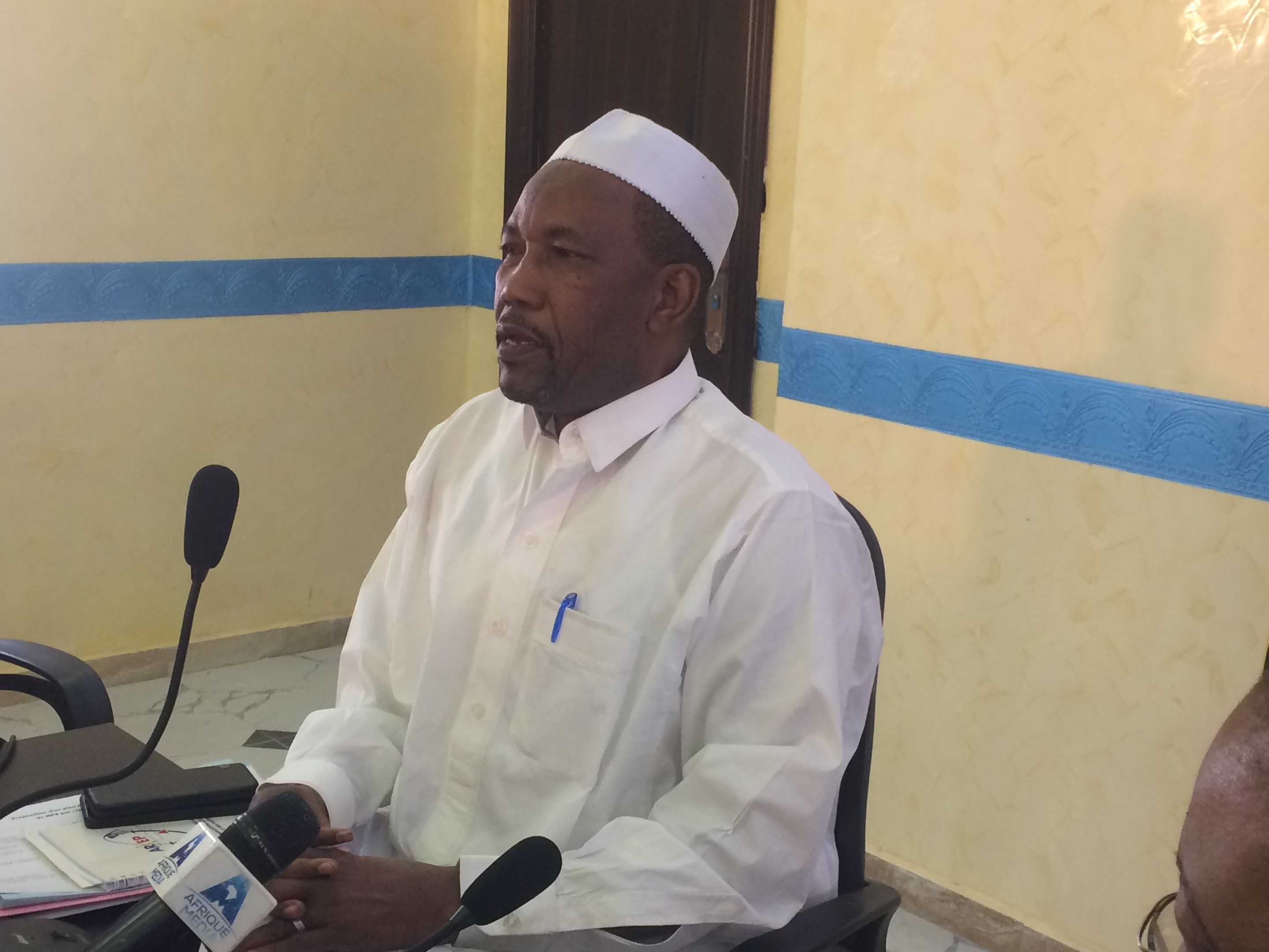 Ein Mann in einem weißen Gewand und einer Mütze sitzt an einem Tisch mit Mikrofonen, während er spricht und Papiere und Gegenstände auf dem Tisch sowie eine andere Person mit Brille rechts von ihm zu sehen sind, in einem Raum mit einer Wand und einer Tür im Hintergrund.