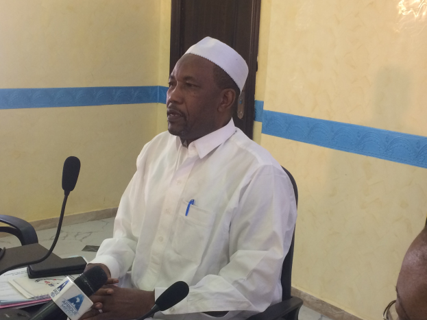 Ein Mann in einem weißen Gewand und einer Mütze sitzt an einem Tisch mit Mikrofonen, während er spricht und Papiere und Gegenstände auf dem Tisch sowie eine andere Person mit Brille rechts von ihm zu sehen sind, in einem Raum mit einer Wand und einer Tür im Hintergrund.